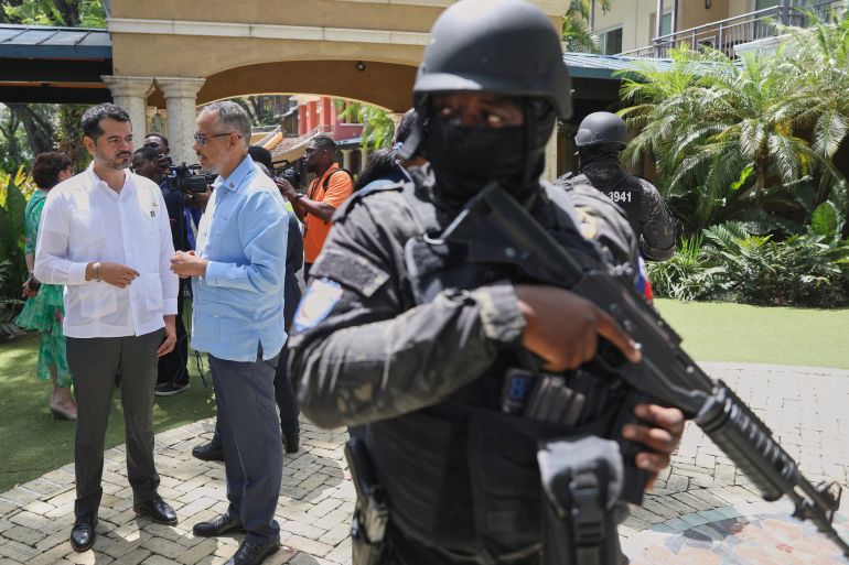 A soldier with a helmet, flak vest and gun stands in front of Haiti's Prime Minister Alix Didier Fils-Aime, center, and Mexico's Charge d'Affaires Jesus Cisneros