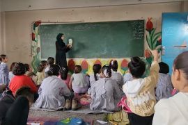 Students sit on the floor as a teacher writes on the board in a classroom.